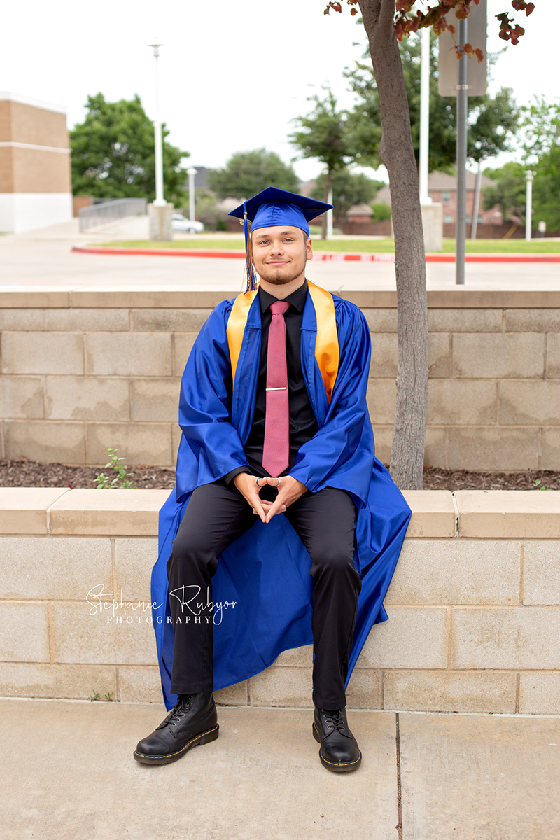 High school senior guy in cap and gown posing for pictures at high school in Fort Worth, Texas. 