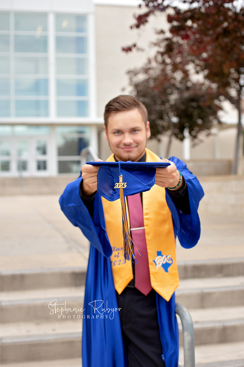 High school senior guy in cap and gown posing for pictures at high school in Fort Worth, Texas. 