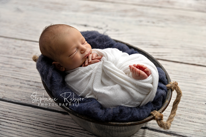 New baby boy posing in a basket wrapped up in white his newborn photo session in Fort Worth.